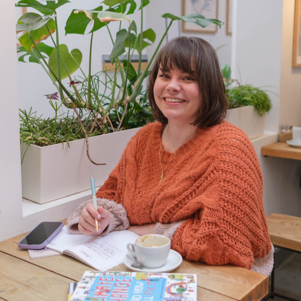 Children's non-fiction author Alice Harman sat at a wooden table in a cafe writing in a notebook with a pen, whilst looking directly at the camera and smiling. There's an empty coffee cup beside her and green plants and grasses in a plat pot beside her.